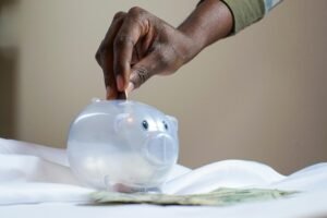 Close-up of a person's hand placing coins into a transparent piggy bank to save money.